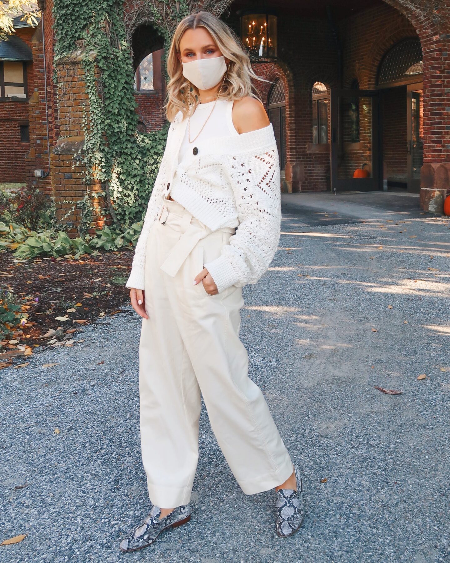 Woman standing in front of an ivy covered building wearing a cream off the shoulder sweater and cream loose fitting trousers to showcase a monochromatic look styled with loafers. 