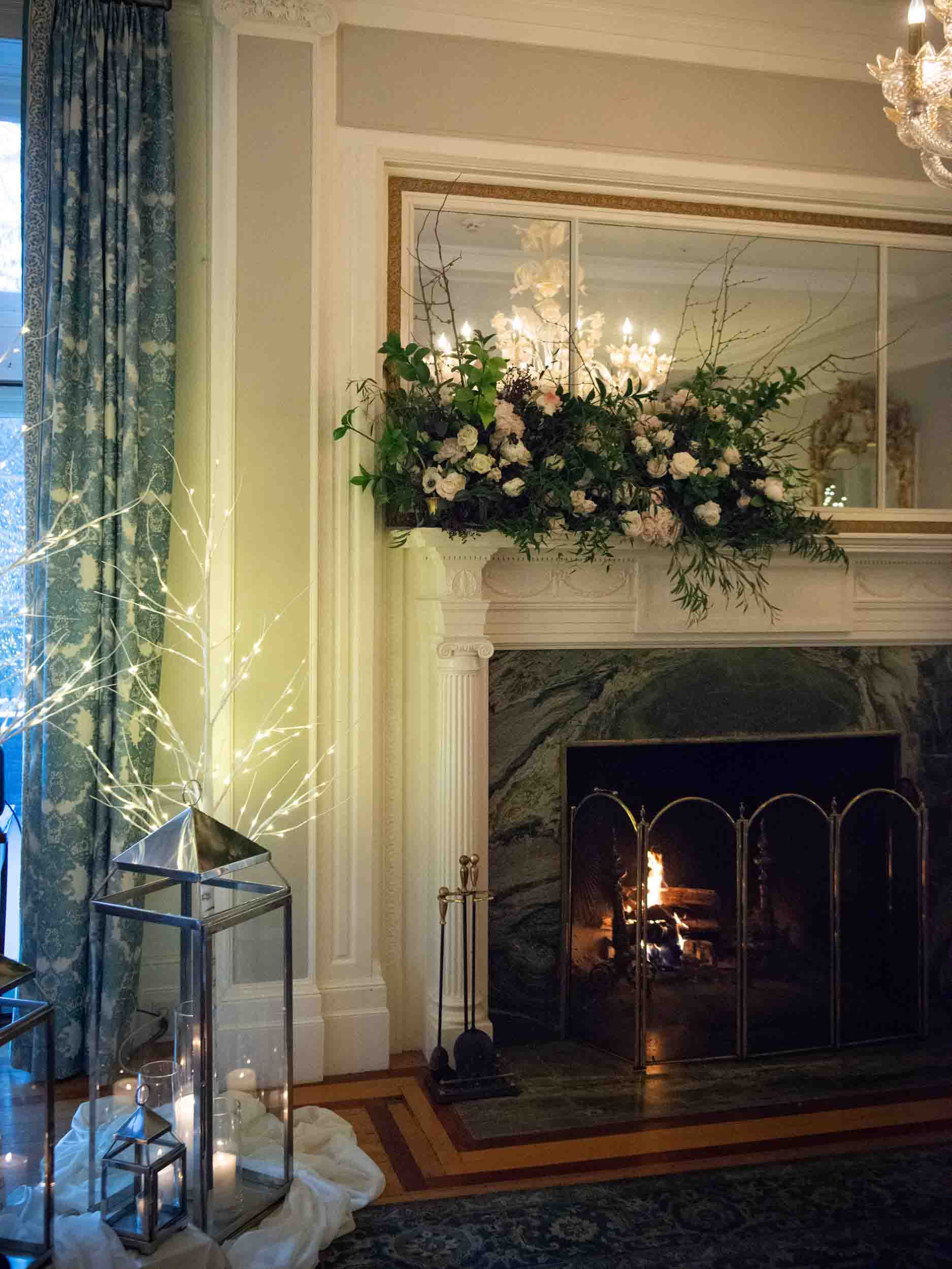 Cluster of white birch trees with sparkly lights, lanterns and candles, next to green marble fireplace with beautiful white, pink and green florals on the mantle